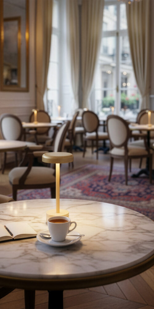 Matt gold IENA cordless table lamp from ELLE Decoration on a marble café table beside a cup of coffee.