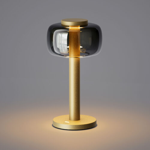 Matt gold ODEON cordless table lamp from ELLE Decoration with an illuminated smoked glass shade, casting warm light onto a grey surface.
