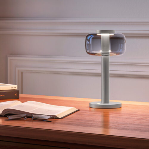 Matt silver ODEON cordless table lamp from ELLE Decoration, illuminated on a wooden desk beside an open book and glasses